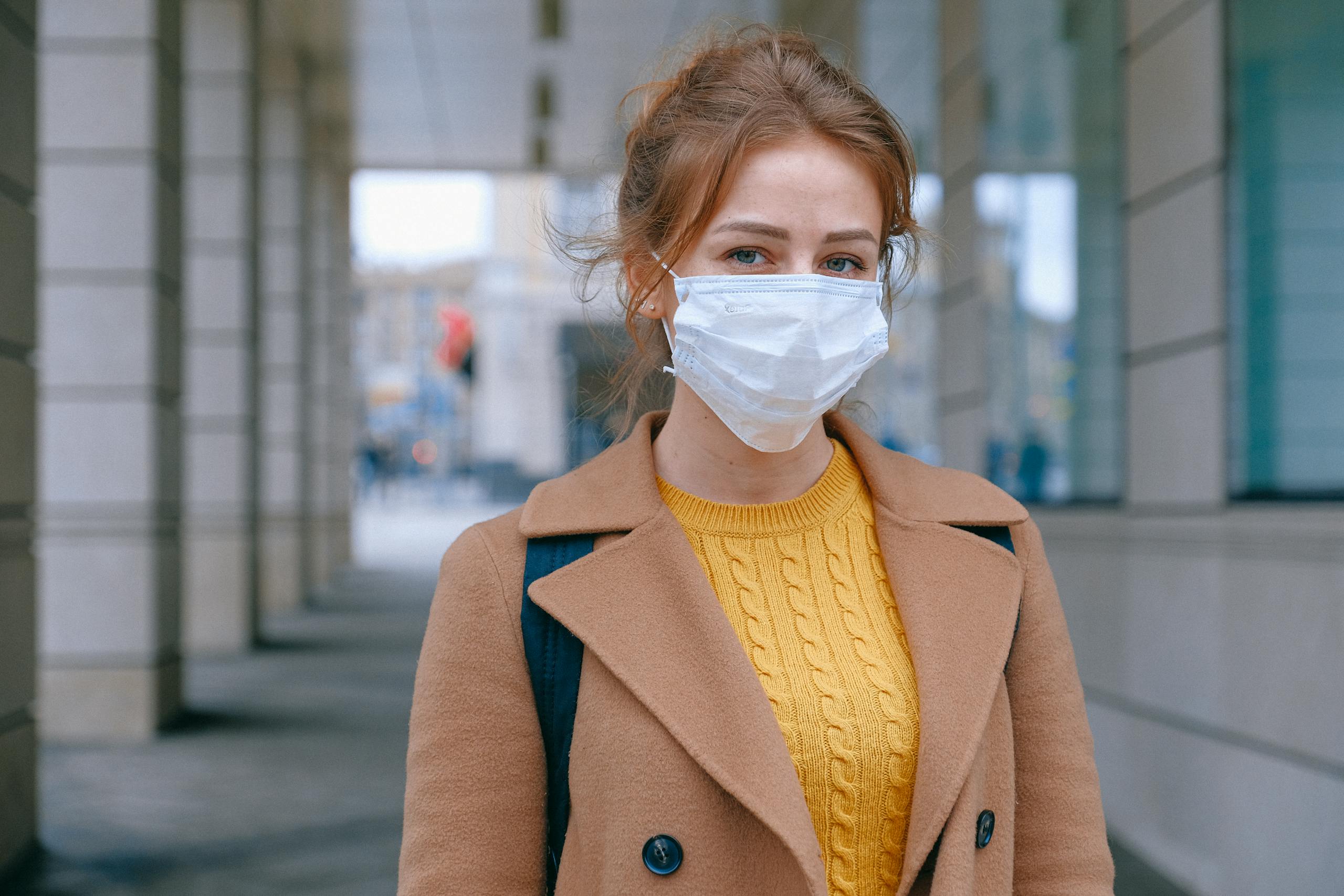 Young woman in a coat and mask stands outdoors, promoting health and safety measures.