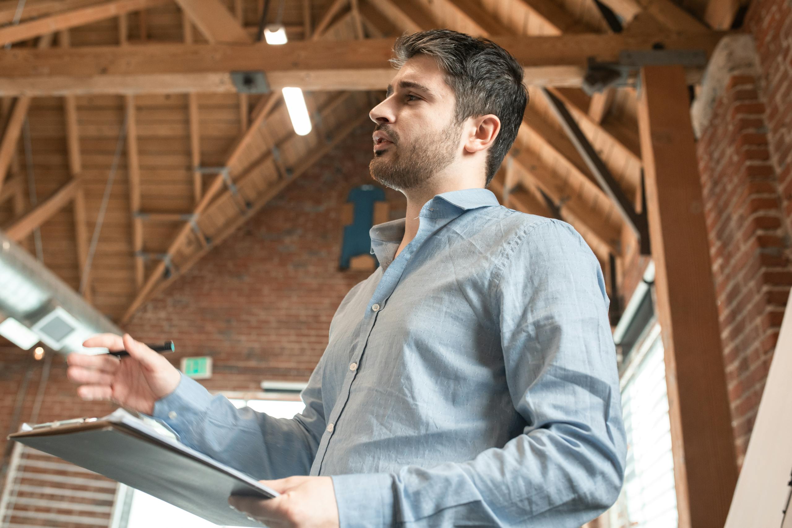 A bearded man in a blue shirt speaking confidently indoors with a clipboard in hand.