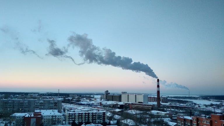 A cityscape featuring industrial smokestacks emitting smoke against a sunset sky, highlighting environmental pollution.