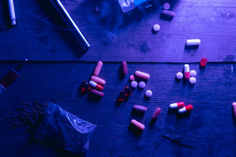 A flat lay of various drugs including pills, capsules, and a syringe on a wooden surface under moody blue lighting.