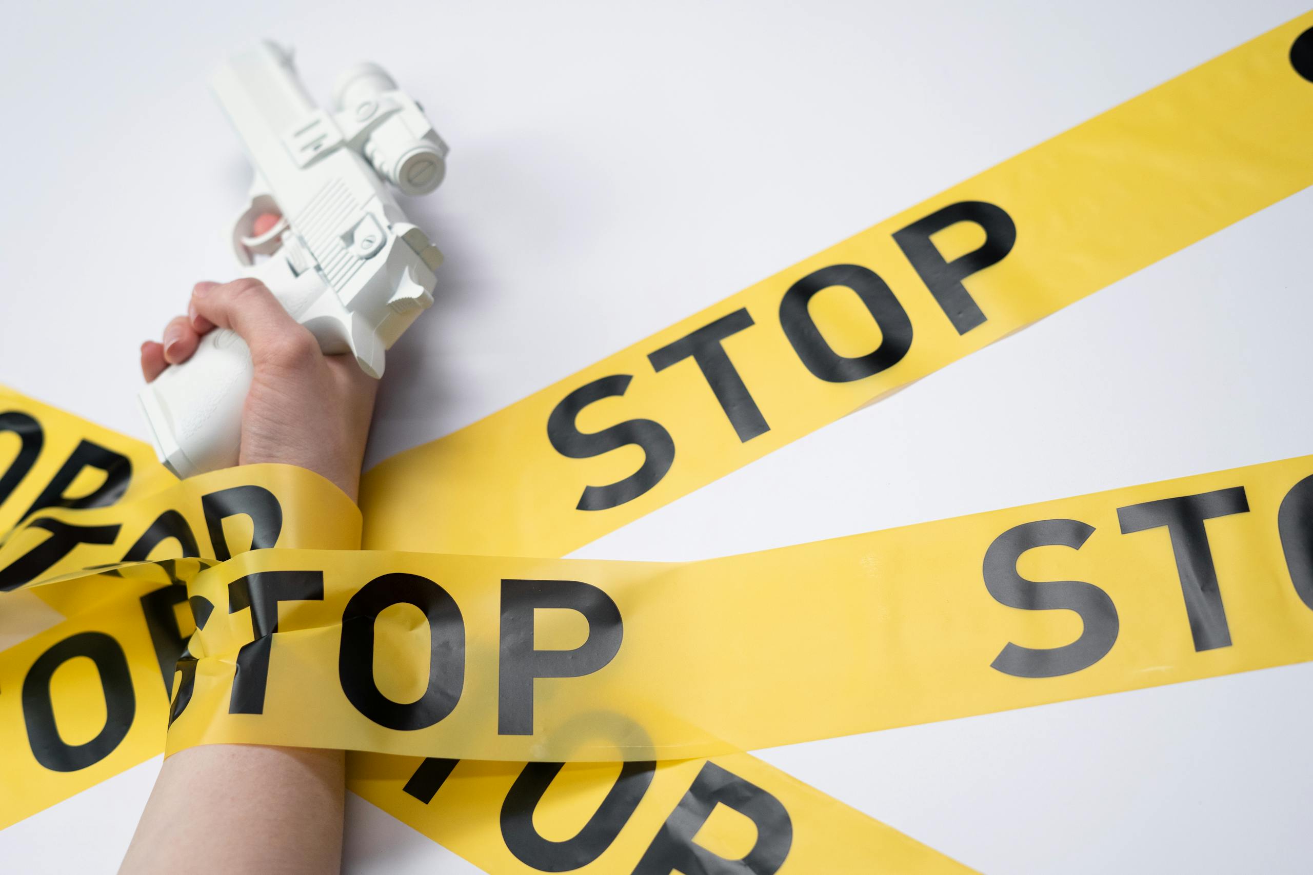 A white toy gun held by a hand wrapped in yellow stop tape on a white background, conveying a message of caution.