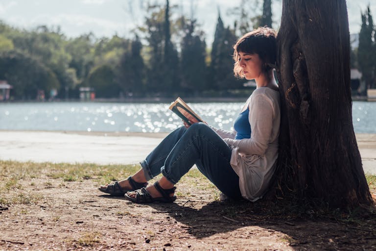 A woman enjoys a peaceful moment reading by the lakeside under a tree in the daytime.