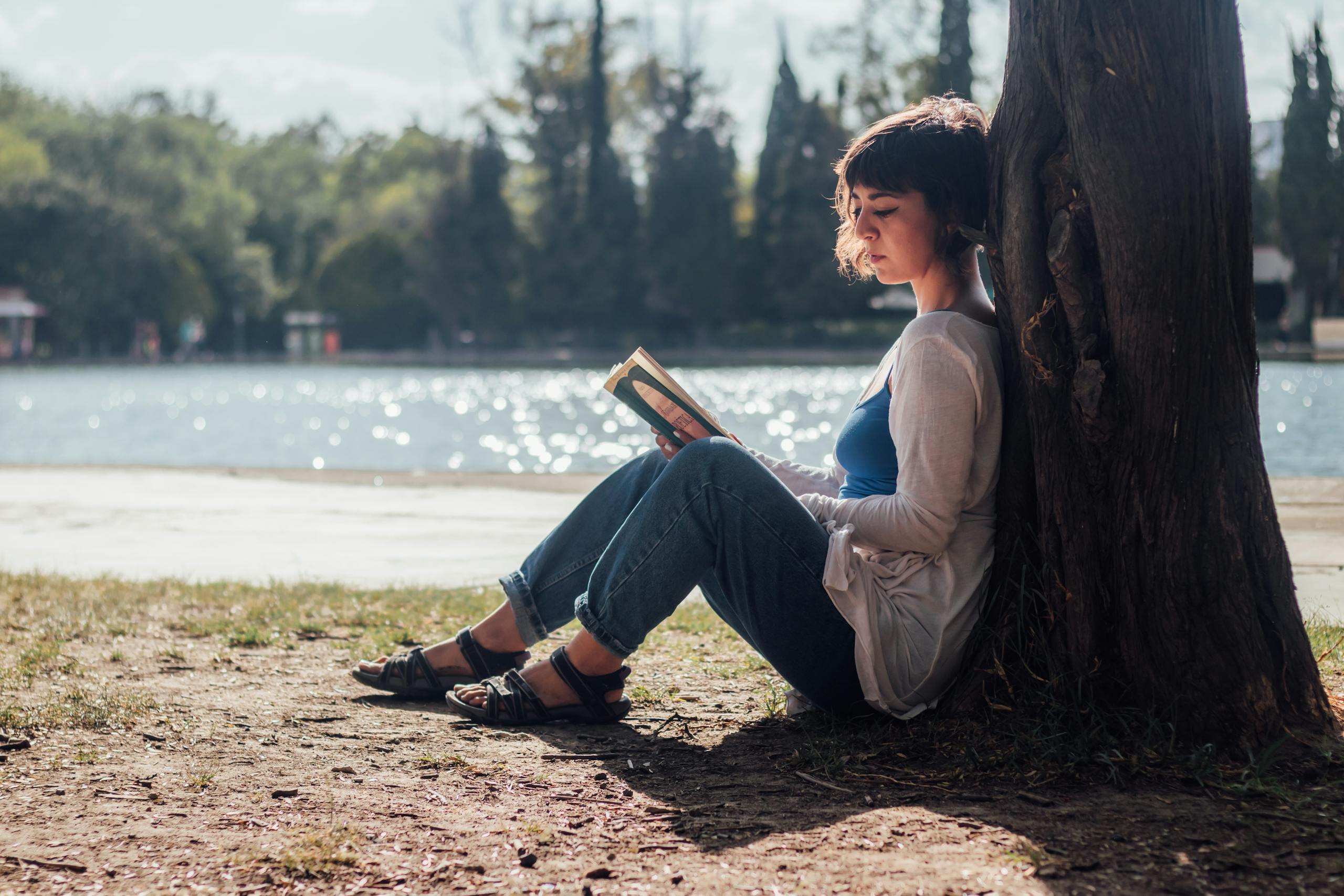 A woman enjoys a peaceful moment reading by the lakeside under a tree in the daytime.