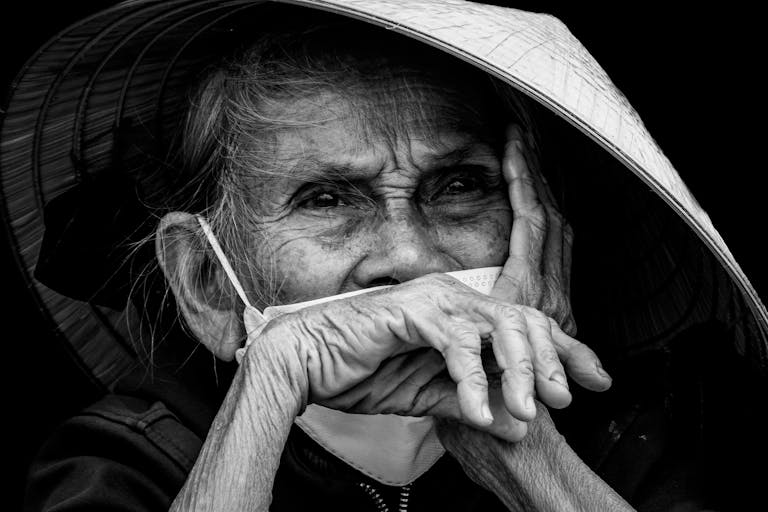 Black and white portrait of an elderly Vietnamese woman wearing a conical hat and mask.