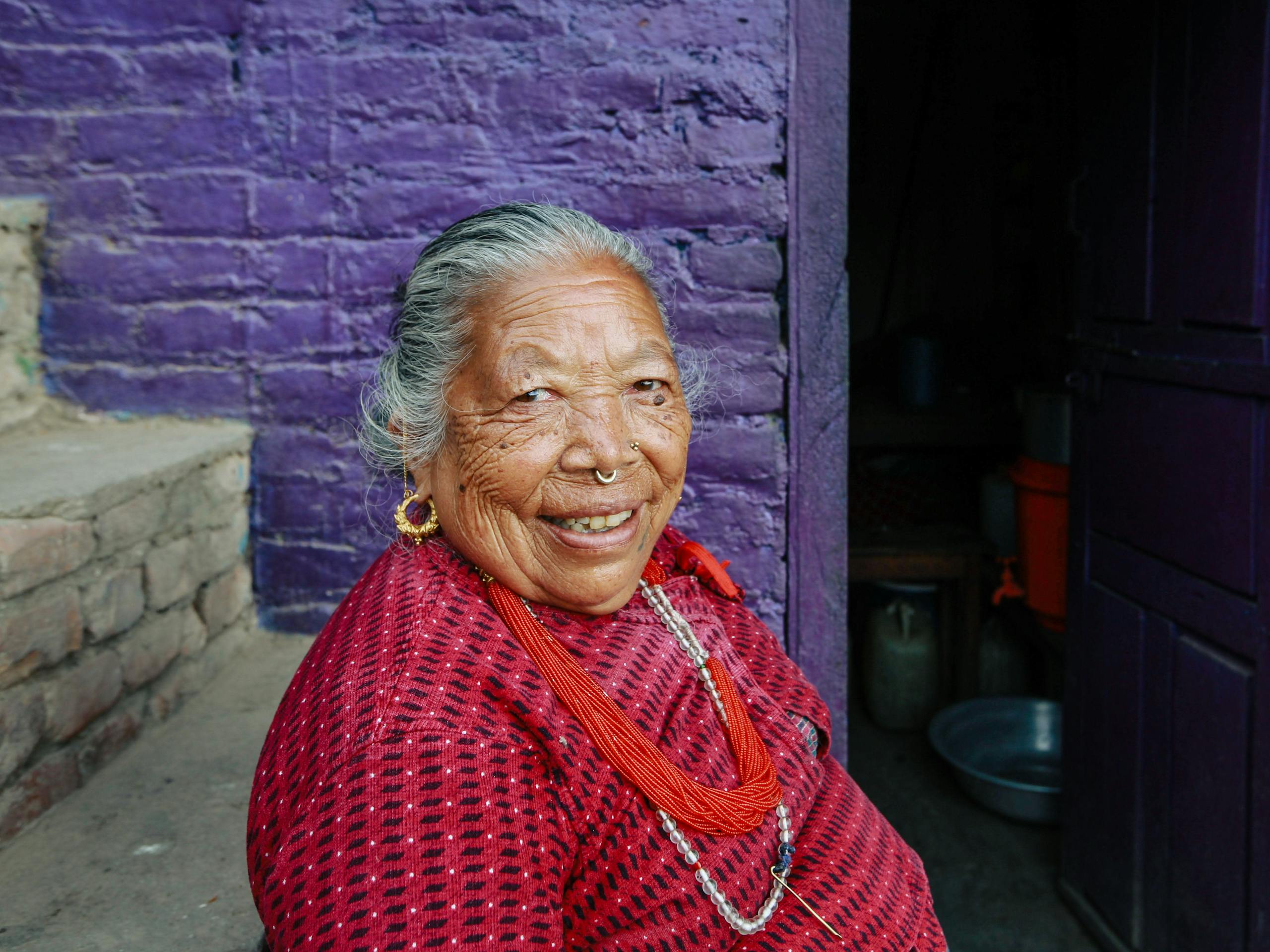 Cheerful elderly woman in traditional attire smiling against a vibrant purple wall.