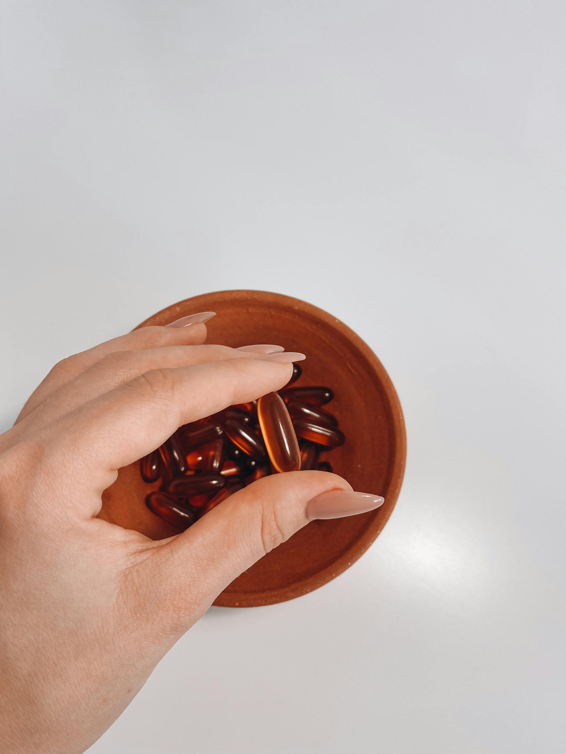 Close-up shot of a hand holding vitamin pills out of a terracotta bowl on a white background.
