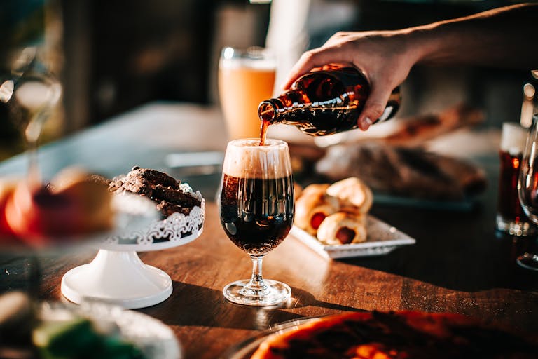 Sunlit scene with hand pouring dark beer into a glass. Includes baked goods and desserts on table.