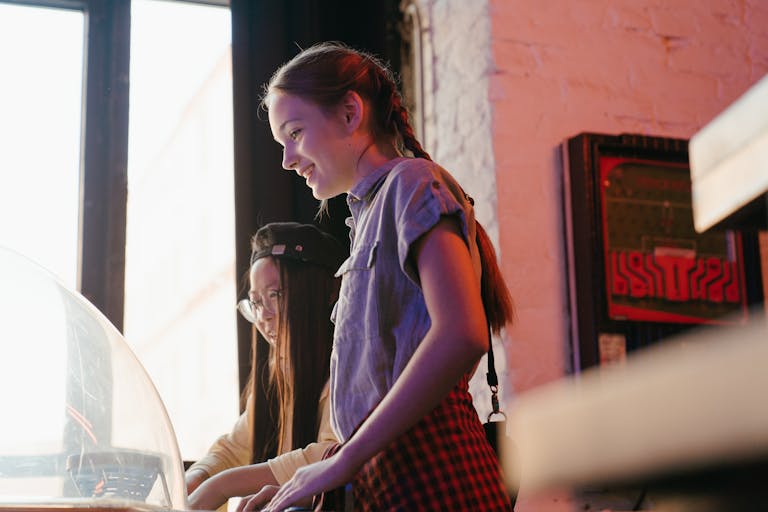 Two teenage girls having fun playing vintage arcade games indoors, highlighting friendship and leisure.