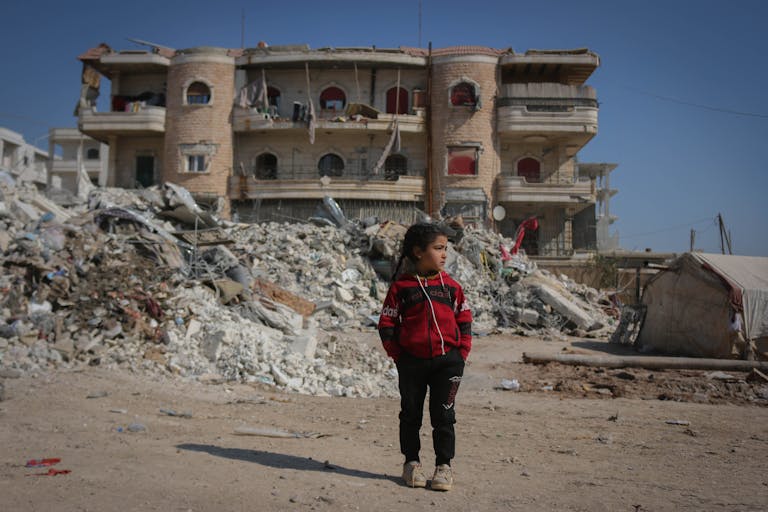 A girl stands in front of a destroyed building in Idlib, Syria, highlighting the impact of conflict.