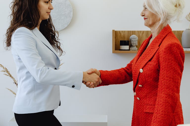 Two professional women elegantly shaking hands in a modern office setting, symbolizing agreement and leadership.